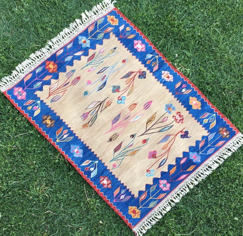 May include: A beige rug with a blue and red border. The rug has a floral pattern with pink, blue, and yellow flowers. The rug has a fringe border.