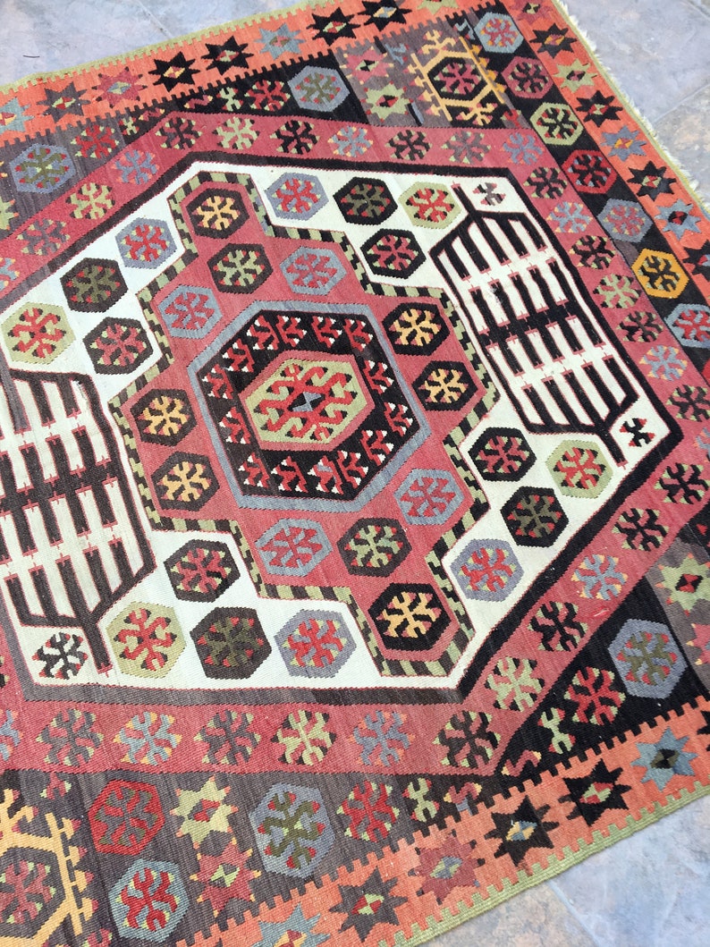 May include: A traditional Turkish rug with a geometric pattern in shades of red, black, white, and green. The rug features a central medallion with a repeating pattern of stars and diamonds.