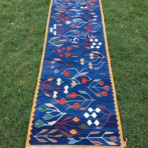 May include: A long, narrow blue rug with a yellow border and white fringe. The rug features a repeating pattern of stylized flowers and leaves in shades of red, orange, yellow, green, and white.
