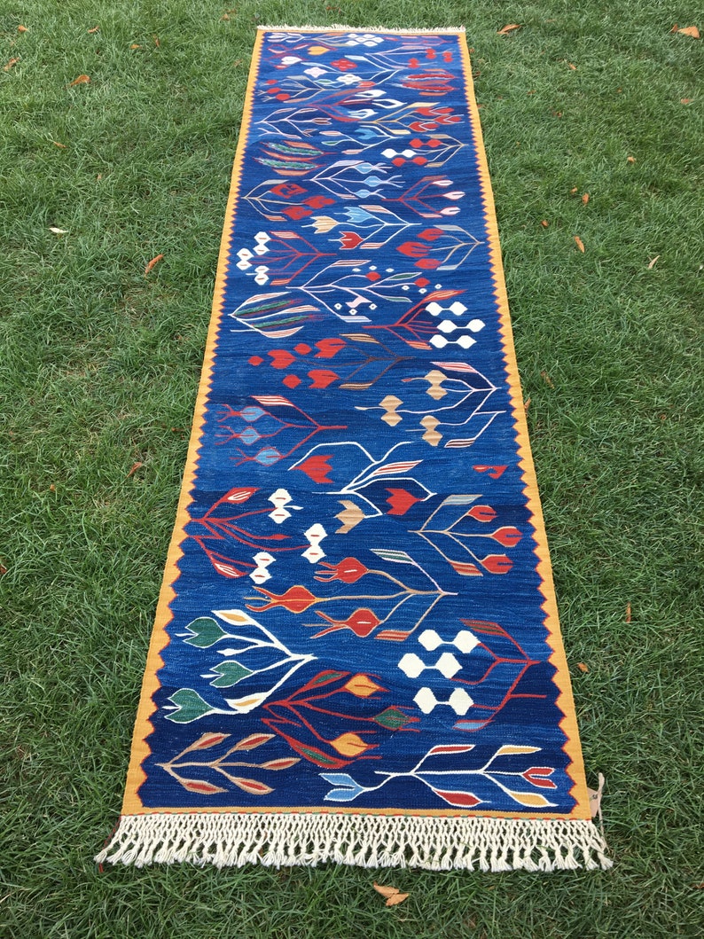 May include: A long, narrow blue rug with a yellow border and white fringe. The rug features a repeating pattern of stylized flowers and leaves in shades of red, orange, yellow, green, and white.