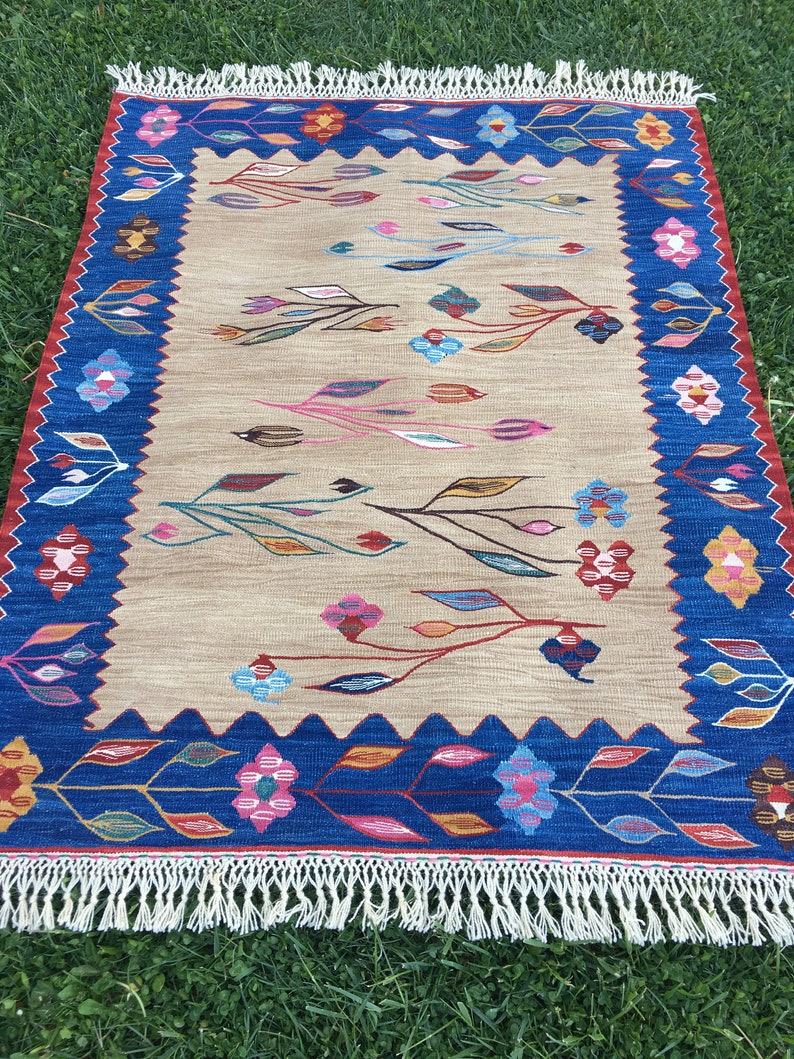May include: A beige rug with a blue border and a floral pattern. The rug has a fringe on all sides.