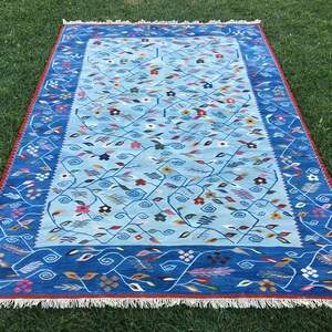 May include: A rectangular rug with a light blue central field featuring a floral pattern of vines and colorful flowers. The border is a darker blue with a repeating vine design. The rug has a red fringe and is displayed on green grass.