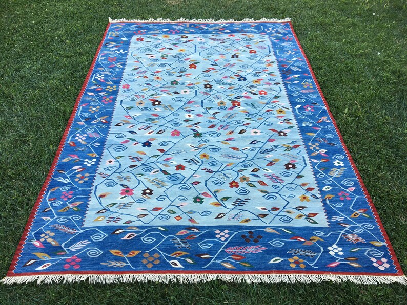 May include: A rectangular rug with a light blue central field featuring a floral pattern of vines and colorful flowers. The border is a darker blue with a repeating vine design. The rug has a red fringe and is displayed on green grass.