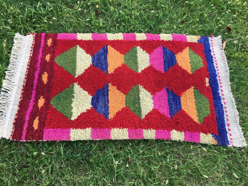 Puede incluir: Una alfombra colorida con un patr&oacute;n geom&eacute;trico de diamantes en tonos de rojo, verde, azul, naranja, rosa y blanco. La alfombra tiene un borde de flecos.