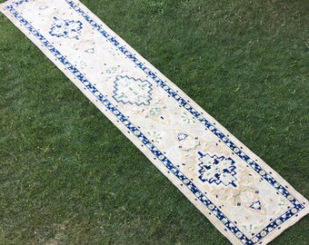May include: A long, cream-colored runner rug with a blue and green floral pattern. The rug has a fringed edge and is laid out on a green grassy surface.