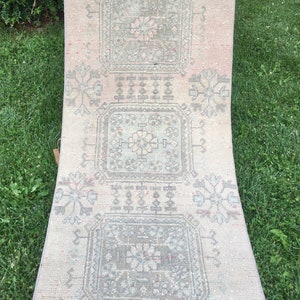 Op de afbeelding: Een lang, rechthoekig tapijt met een witte achtergrond en een herhalend patroon van grijze en witte geometrische vormen. Het tapijt heeft een verweerde, vintage uitstraling.
