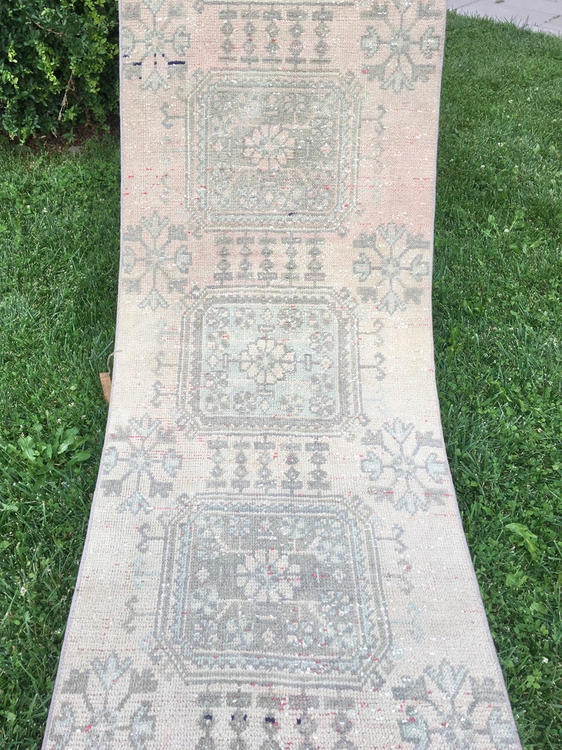 Op de afbeelding: Een lang, rechthoekig tapijt met een witte achtergrond en een herhalend patroon van grijze en witte geometrische vormen. Het tapijt heeft een verweerde, vintage uitstraling.