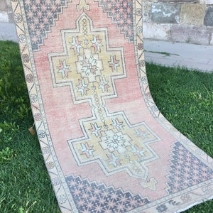 May include: A rectangular, vintage-style rug with a central geometric design in yellow and white, set against a faded pink background. The borders feature repeating floral and geometric patterns in muted tones of beige, gray, and pink. The rug is displayed on green grass.