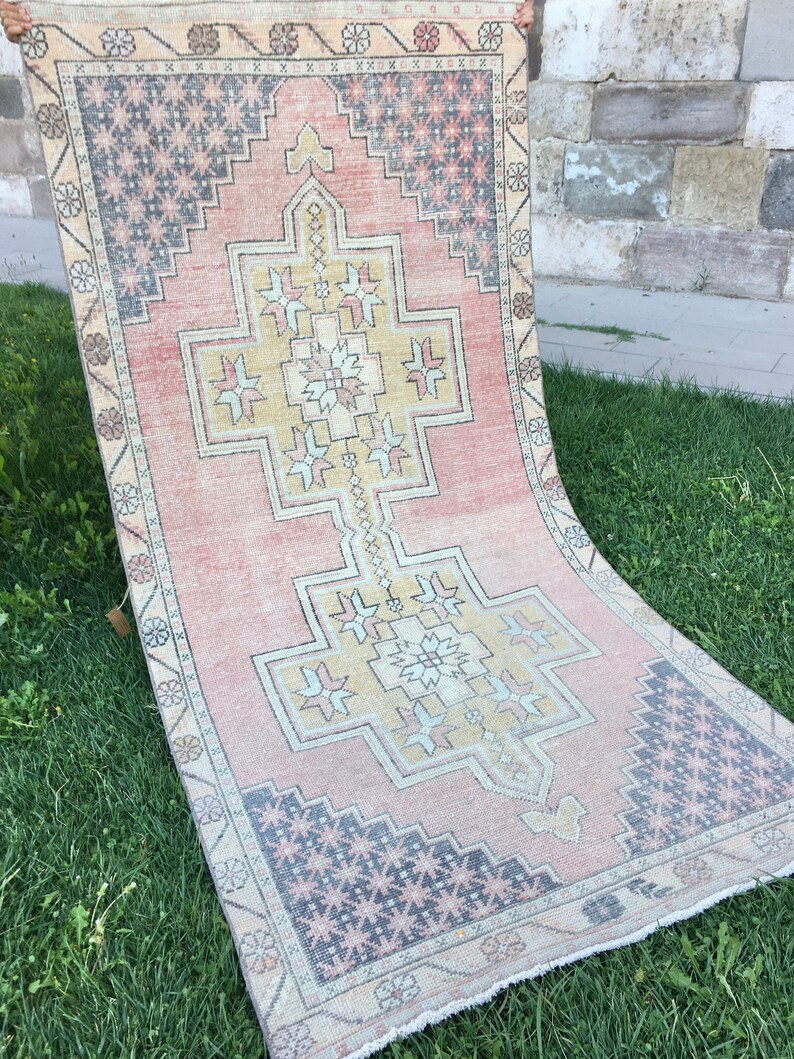 May include: A rectangular, vintage-style rug with a central geometric design in yellow and white, set against a faded pink background. The borders feature repeating floral and geometric patterns in muted tones of beige, gray, and pink. The rug is displayed on green grass.