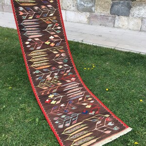 Puede incluir: Una alfombra larga y estrecha con fondo marr&oacute;n y un patr&oacute;n floral colorido. La alfombra tiene un borde rojo y flecos blancos.