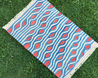 May include: A blue and red patterned rug with a white background and fringe on all sides. The rug has a wavy design with red diamonds.