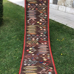 Puede incluir: Una alfombra larga y estrecha con fondo marr&oacute;n y un patr&oacute;n repetitivo de flores y hojas estilizadas en tonos de rojo, verde, azul, amarillo y rosa. La alfombra tiene un borde rojo y flecos blancos.