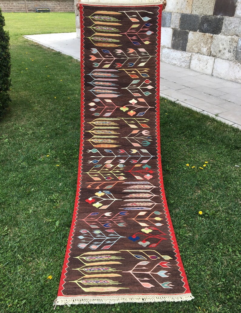 Puede incluir: Una alfombra larga y estrecha con fondo marr&oacute;n y un patr&oacute;n repetitivo de flores y hojas estilizadas en tonos de rojo, verde, azul, amarillo y rosa. La alfombra tiene un borde rojo y flecos blancos.
