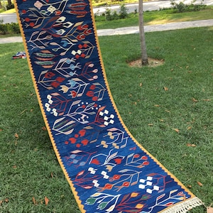 May include: A long, narrow blue rug with a yellow border and white fringe. The rug features a repeating pattern of stylized flowers and leaves in shades of red, white, blue, green, and yellow.