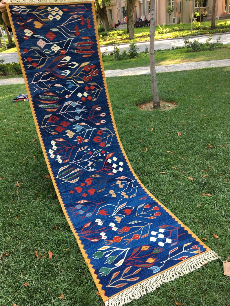 May include: A long, narrow blue rug with a yellow border and white fringe. The rug features a repeating pattern of stylized flowers and leaves in shades of red, white, blue, green, and yellow.