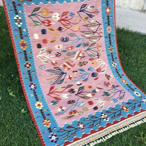 Op de afbeelding: Een traditioneel Turks tapijt met een roze achtergrond en een blauwe rand. Het tapijt heeft een bloemenpatroon met ingewikkelde details. Het tapijt is geweven van wol en heeft franjes aan de randen.