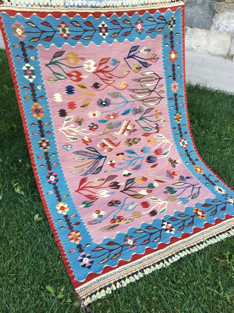 Op de afbeelding: Een traditioneel Turks tapijt met een roze achtergrond en een blauwe rand. Het tapijt heeft een bloemenpatroon met ingewikkelde details. Het tapijt is geweven van wol en heeft franjes aan de randen.