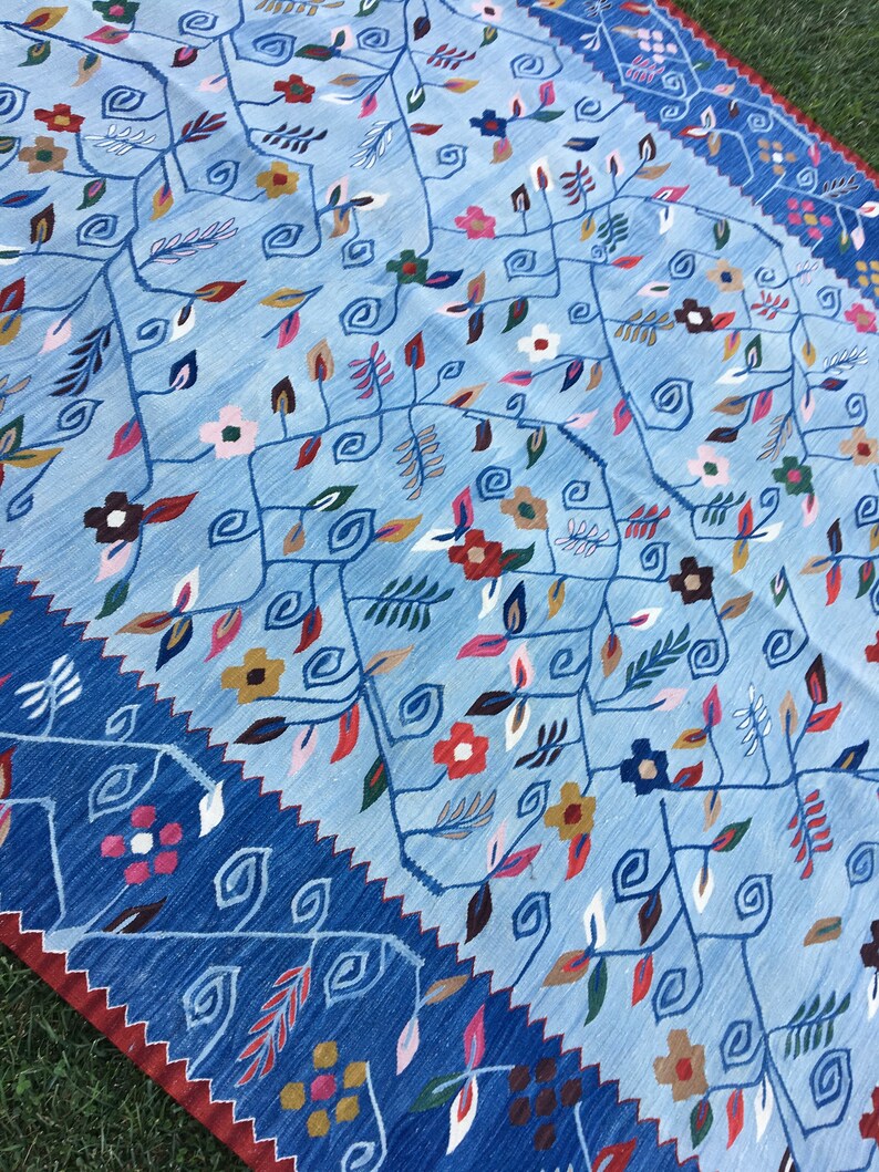 May include: A light blue rug with a floral pattern. The rug features a central design of stylized vines and flowers in shades of red, pink, yellow, and brown. A dark blue border with a zigzag pattern frames the rug.
