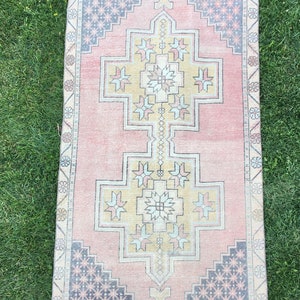 May include: A rectangular, vintage-style rug with a central geometric design in shades of pink, yellow, and blue. The rug features a repeating pattern of stars and floral motifs, with a faded, distressed appearance. The rug is placed on a green grassy surface.