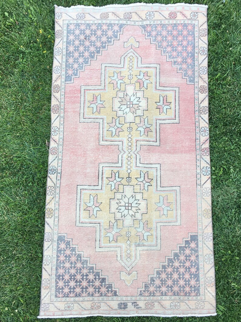 May include: A rectangular, vintage-style rug with a central geometric design in shades of pink, yellow, and blue. The rug features a repeating pattern of stars and floral motifs, with a faded, distressed appearance. The rug is placed on a green grassy surface.