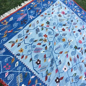 May include: A blue and light blue patterned rug with floral and leaf designs. The rug has a dark blue border with a red edge and white fringe. The rug is made of woven fabric and is placed on green grass.