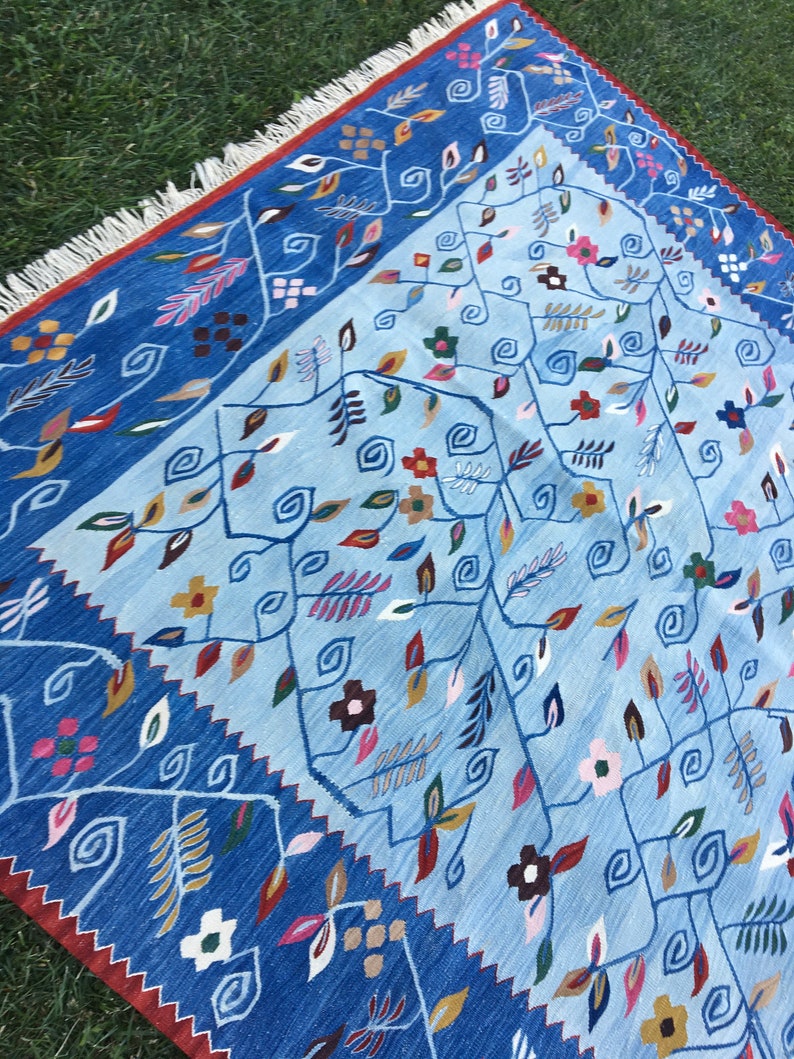 May include: A blue and light blue patterned rug with floral and leaf designs. The rug has a dark blue border with a red edge and white fringe. The rug is made of woven fabric and is placed on green grass.