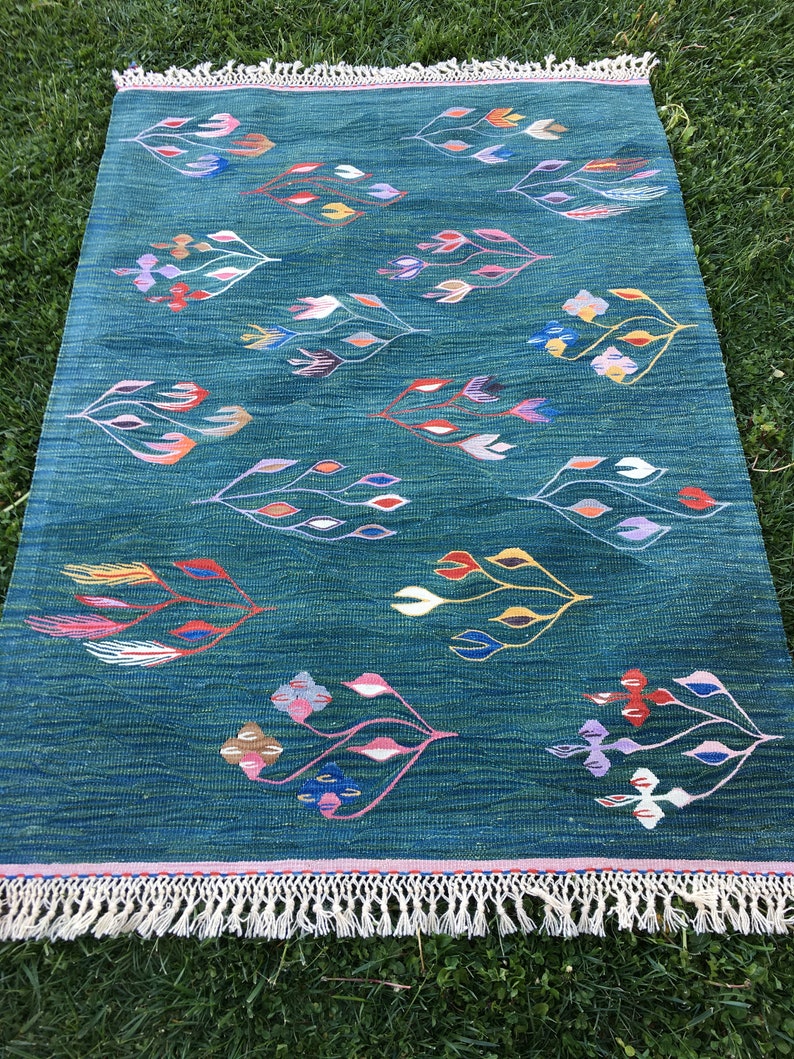 Puede incluir: Una alfombra azul turquesa con un patr&oacute;n geom&eacute;trico de hojas estilizadas de colores. La alfombra tiene un borde de flecos blancos.