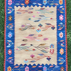 May include: A beige rug with a blue and red border. The rug has a floral pattern with colorful leaves and flowers.