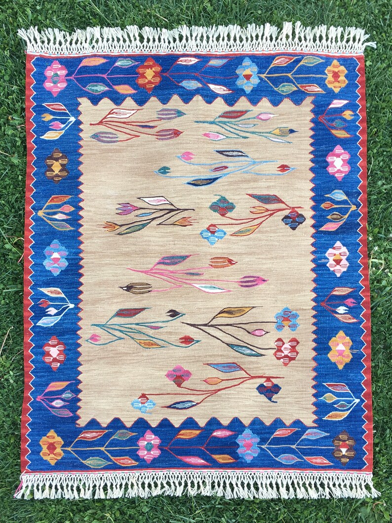 May include: A beige rug with a blue and red border. The rug has a floral pattern with colorful leaves and flowers.