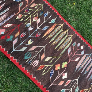 Puede incluir: Una alfombra de color marr&oacute;n y rojo con un dise&ntilde;o floral. La alfombra tiene un patr&oacute;n geom&eacute;trico con flores y hojas estilizadas en tonos de rojo, azul, amarillo, verde y rosa.