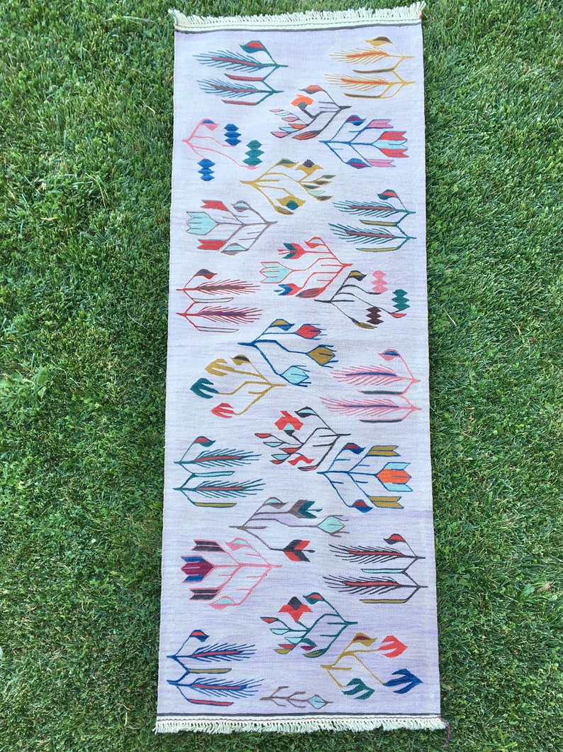 May include: A long, rectangular rug with a white background and a colorful, abstract pattern of stylized leaves and flowers. The rug has a fringe border.