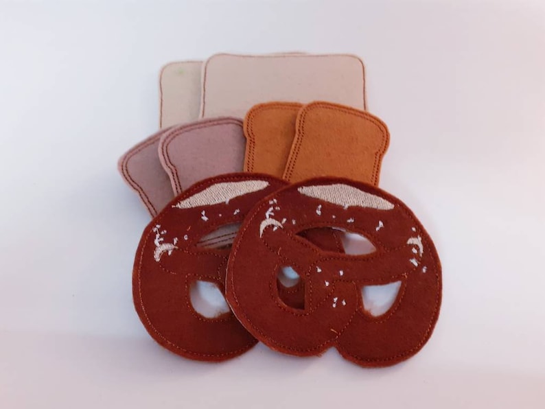Puede incluir: Dos formas de pretzel de fieltro marr&oacute;n con bordados blancos, y tres formas de pan de fieltro beige, marr&oacute;n claro y marr&oacute;n oscuro.