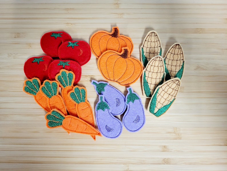 Puede incluir: Apliques de fieltro de varias verduras y frutas, incluyendo tomates rojos, calabazas naranjas, zanahorias verdes, berenjenas moradas y ma&iacute;z amarillo.