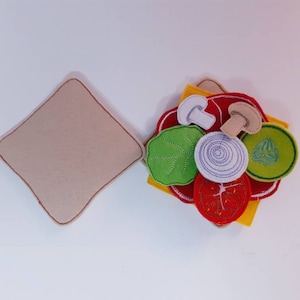 Puede incluir: Un juego de comida de fieltro para jugar con una rebanada de pan marr&oacute;n y varios ingredientes, incluyendo rodajas de tomate rojo, lechuga verde, un hongo blanco, una cebolla blanca y una rodaja de pepino verde.