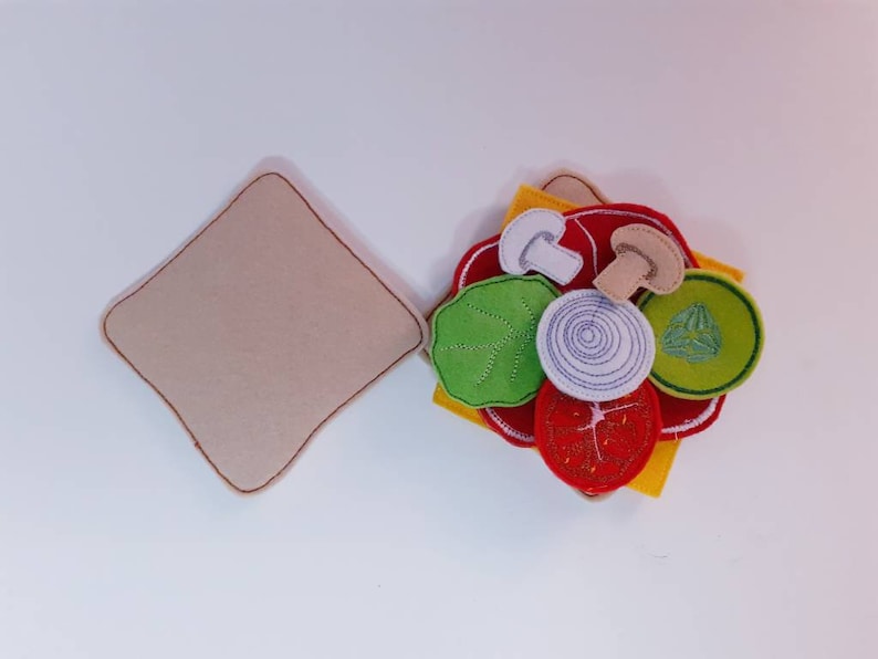 Puede incluir: Un juego de comida de fieltro para jugar con una rebanada de pan marr&oacute;n y varios ingredientes, incluyendo rodajas de tomate rojo, lechuga verde, un hongo blanco, una cebolla blanca y una rodaja de pepino verde.