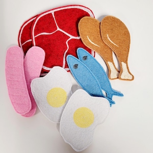 Puede incluir: Conjunto de comida de fieltro con un filete rojo, dos patas de pollo marrones, dos peces azules, dos huevos blancos con yemas amarillas y una salchicha rosa.