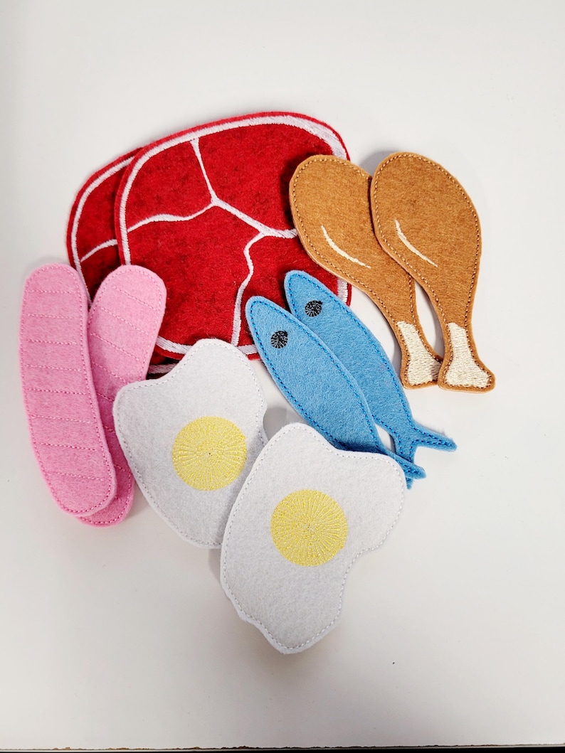 Puede incluir: Conjunto de comida de fieltro con un filete rojo, dos patas de pollo marrones, dos peces azules, dos huevos blancos con yemas amarillas y una salchicha rosa.