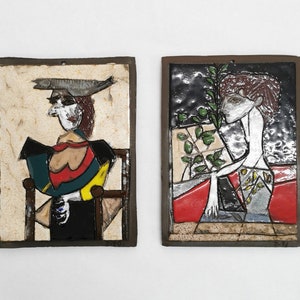 Peut inclure: Deux plaques murales en céramique avec des figures abstraites. La plaque de gauche représente une figure avec un poisson sur la tête, portant une tenue rouge, jaune et verte. La plaque de droite représente une figure avec une plante derrière elle, portant une tenue grise et rouge.