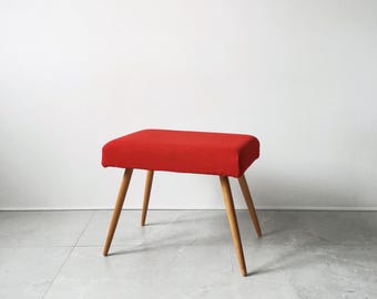 May include: A small, red upholstered stool with four wooden legs. The stool has a rectangular seat and a simple, mid-century modern design. The legs are angled outwards for stability.