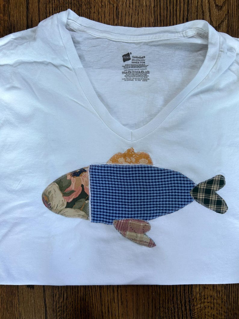 May include: White V-neck t-shirt with a colorful fish applique. The fish design features floral, blue gingham, and plaid fabric patches. The fish has a yellow-orange dorsal fin and a plaid tail fin. The shirt is made of a soft, comfortable material.