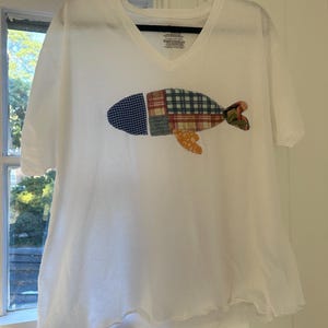 Op de afbeelding: Wit V-hals T-shirt met een patchwork visontwerp. De vis heeft blauwe, groene, rode en oranje stoffen patronen. Het shirt heeft korte mouwen en een licht gerimpelde zoom. Het shirt hangt aan een zwarte hanger.