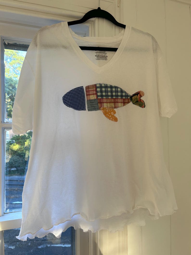 Puede incluir: Camiseta blanca con cuello en V con un dise&ntilde;o de pez patchwork. El pez presenta tela estampada azul, verde, roja y naranja. La camiseta tiene mangas cortas y un dobladillo ligeramente con volantes. La camiseta est&aacute; colgada en una percha negra.