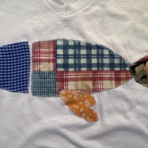 Puede incluir: Camiseta blanca con cuello en V con un dise&ntilde;o de pez patchwork. El pez est&aacute; compuesto por varios patrones de tela, incluyendo cuadros azules, cuadros y estampados florales. La cola y las aletas del pez son en tonos rosa, naranja y verde.