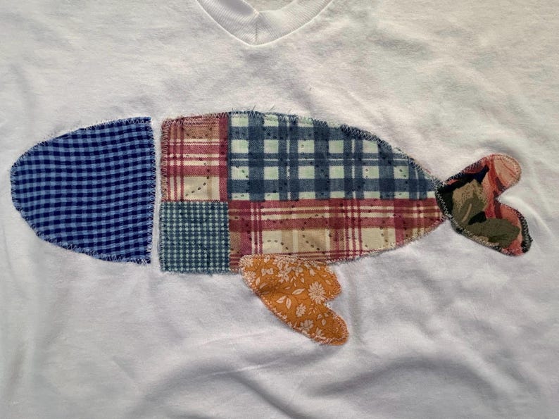 Puede incluir: Camiseta blanca con cuello en V con un dise&ntilde;o de pez patchwork. El pez est&aacute; compuesto por varios patrones de tela, incluyendo cuadros azules, cuadros y estampados florales. La cola y las aletas del pez son en tonos rosa, naranja y verde.