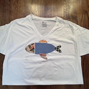 May include: White V-neck t-shirt with a patchwork fish design. The fish features a floral head, a blue gingham body, and a plaid tail and fins. The shirt is made of a soft, comfortable material.
