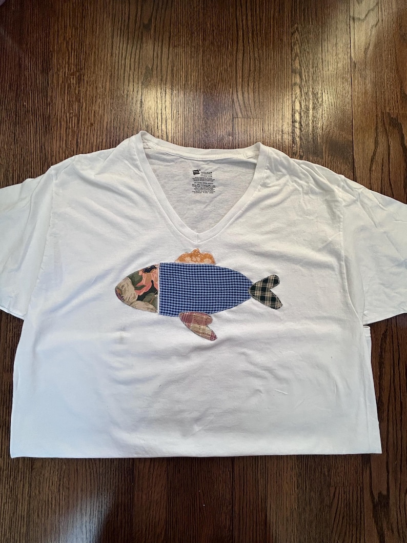 May include: White V-neck t-shirt with a patchwork fish design. The fish features a floral head, a blue gingham body, and a plaid tail and fins. The shirt is made of a soft, comfortable material.