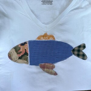May include: White V-neck t-shirt with a patchwork fish design. The fish features a floral print, blue gingham, and plaid patterns in shades of brown, green, and blue. The fish's fin is a golden-orange color.
