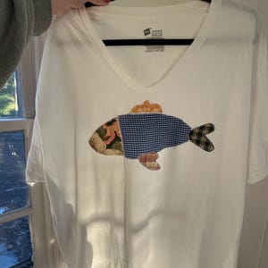 May include: White V-neck t-shirt with a patchwork fish design. The fish features a mix of patterned fabrics in shades of blue, green, orange, and floral prints. The shirt is held up by a black hanger.