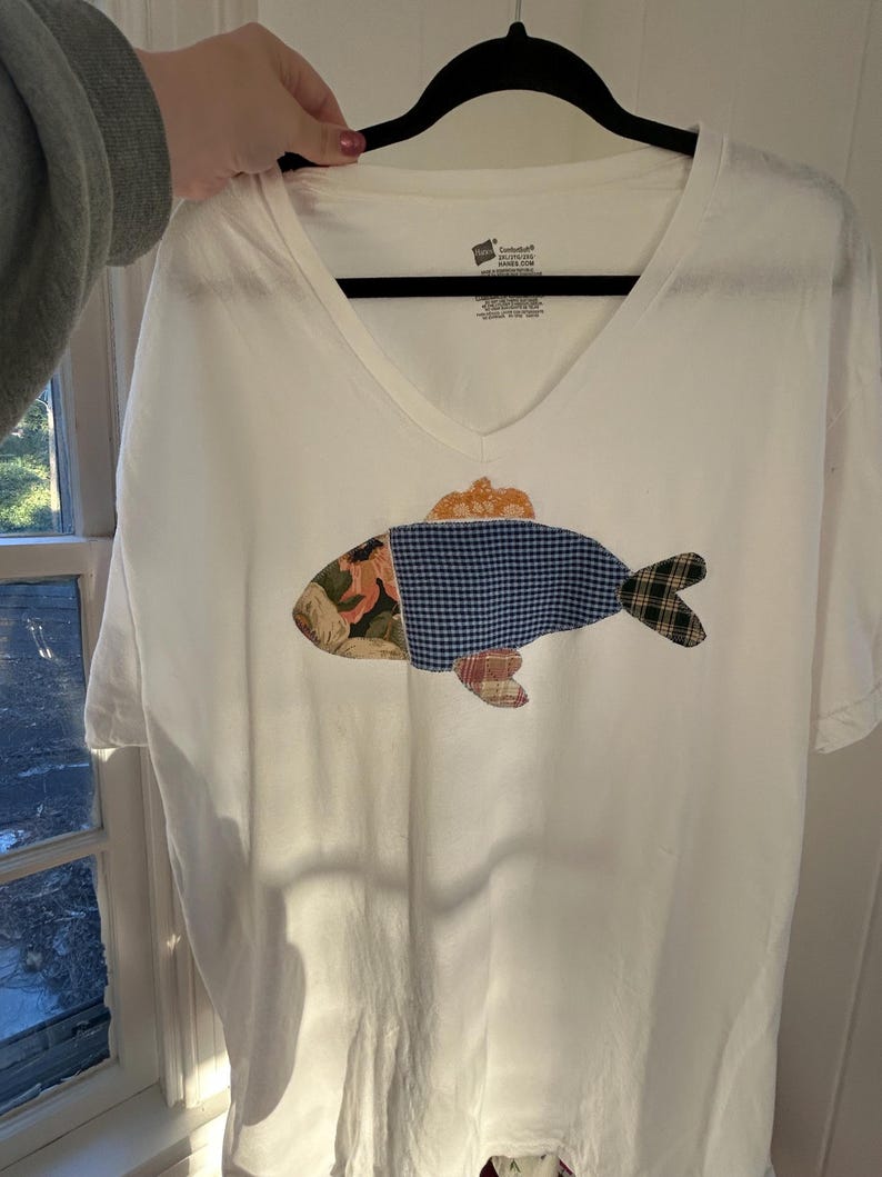 May include: White V-neck t-shirt with a patchwork fish design. The fish features a mix of patterned fabrics in shades of blue, green, orange, and floral prints. The shirt is held up by a black hanger.
