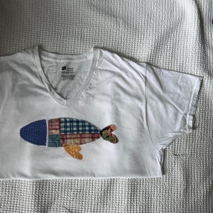 Könnte beinhalten: Ein weißes T-Shirt mit V-Ausschnitt und einer mittigen Applikation in Fischform. Der Fisch besteht aus verschiedenen Stoffstücken, darunter blaues Vichy-Karo, rot-blaues Schottenkaro und ein gelber Blumendruck, was ein strukturiertes Design ergibt.